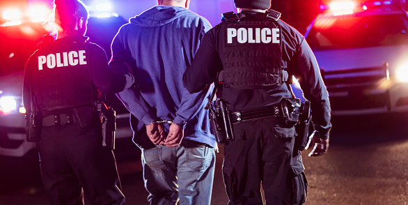 Cropped rear view of two multiracial police officers arresting a criminal suspect, a man with his hands handcuffed behind his back. One of the officers is an African-American man and the other is a policewoman, both in their 40s.