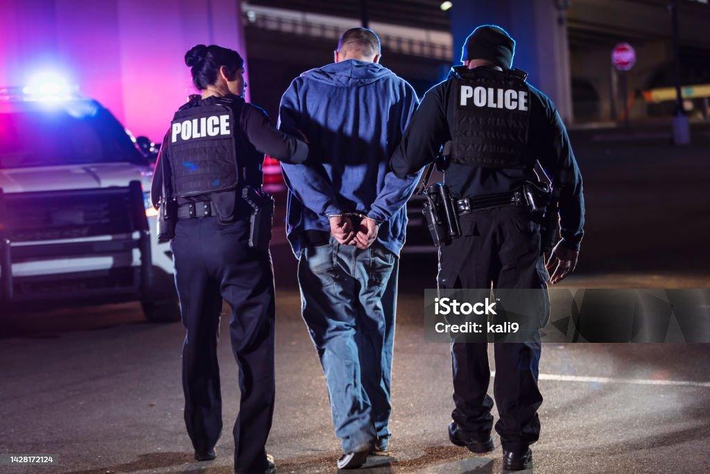 Handcuffed suspect being arrested by police at night Rear view of two multiracial police officers arresting a criminal suspect, a man with his hands handcuffed behind his back. One of the officers is an African-American man and the other is a policewoman, both in their 40s. Arrest Stock Photo