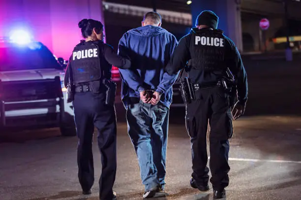 Rear view of two multiracial police officers arresting a criminal suspect, a man with his hands handcuffed behind his back. One of the officers is an African-American man and the other is a policewoman, both in their 40s.