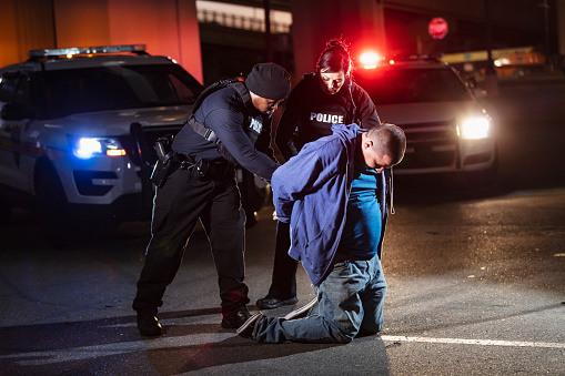 Two multiracial police officers arresting a suspect who is kneeling in a parking lot with his hands behind his back. It is nighttime and the emergency lights and headlights of the police cars are on behind them. The African-American male officer is handcuffing the suspect, a man in his 30s. The other officer is a policewoman.