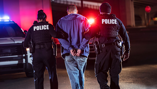 Rear view of two multiracial police officers arresting a criminal suspect, a man with his hands handcuffed behind his back. One of the officers is an African-American man and the other is a policewoman, both in their 40s.