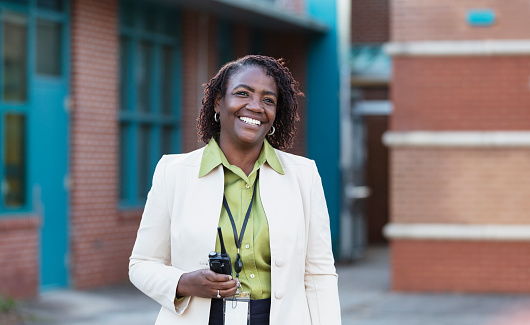 A mature African-American woman standing outside a school building, smiling and looking away from the camera. She is a teacher or school principal.