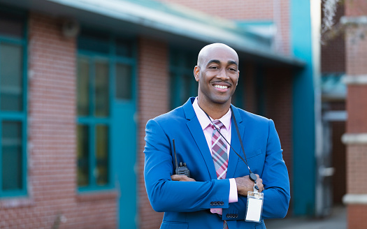 A mature African-American man wearing a suit and tie, standing in front of a school building. He is the school principal, administrator or teacher, smiling confidently at the camera.
