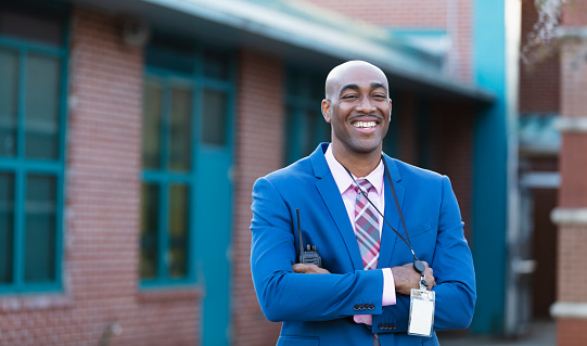 A mature African-American man wearing a suit and tie, standing in front of a school building. He is the school principal, administrator or teacher, smiling confidently at the camera.