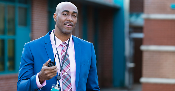 A mature African-American man wearing a suit and tie, standing in front of a school building with a walkie-talkie. He is the school principal, administrator or teacher.