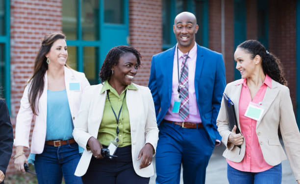 A multiracial group of four teachers, three women and one man, walking together outside a school building, smiling. They are wearing id tags, and the African-American woman is carrying a walkie-talkie.