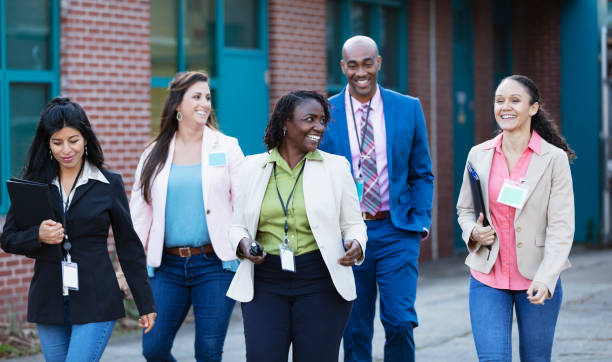 Group of teachers walking together outside school A multiracial group of five teachers, four women and one man, walking together outside a school building, smiling. They are wearing id tags, and the African-American woman in the middle is carrying a walkie-talkie. school administrator stock pictures, royalty-free photos & images