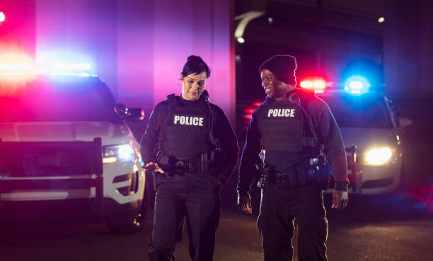 Two police officers working at night, conversing Two multiracial police officers working at night, walking side by side, conversing and smiling, their police cars behind them with emergency lights and headlights on. One of the officers is an African-American man and the other is a policewoman, both in their 40s. police force smiling officer stock pictures, royalty-free photos & images