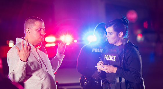 Two multiracial police officers working at night, taking a statement from a victim or witness to a crime. It is nighttime, and the emergency lights of a police car are visible in the background.  The focus is on the policewoman and the civilian in the foreground. The other officer is an African-American man.