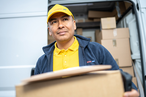 Mid adult courier in uniform carrying cardboard box, looking up. Smiling delivery man unloading truck. Shipment service, postal worker holding customer order