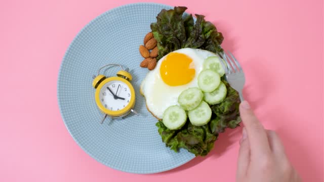 Hand puts an alarm clock on plate of food on pink background, concept of intermittent fasting.