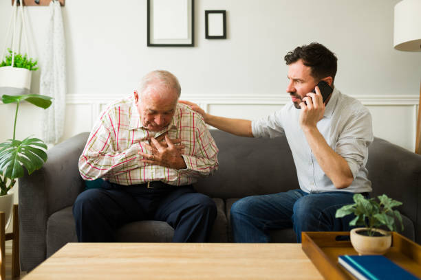 Concerned son calling emergency 911 due to a heart attack of his elderly father Sad man taking care of his father with health problems at home while having a heart attack and calling on the phone emergency medical services chest pain calling doctor stock pictures, royalty-free photos & images