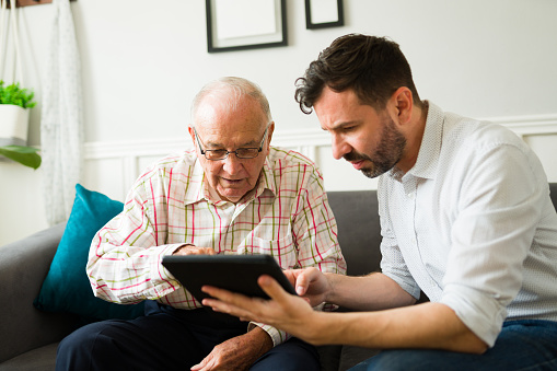 Senior elderly man asking questions about how to use a digital tablet and technology to his caucasian son