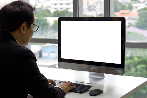 Asian senior manager in black suit sit at desk in office typing on computer keyboard with white screen display.