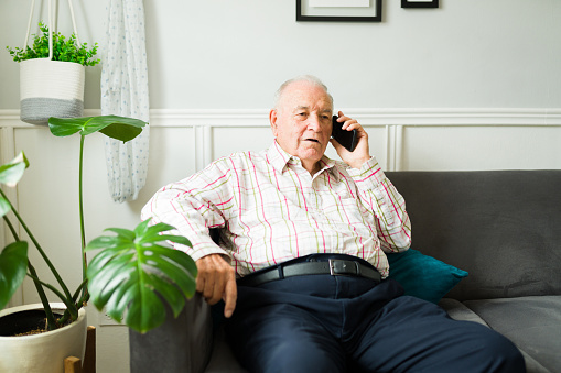 Caucasian old man looking relaxed sitting and resting on the sofa while talking on the phone with family or friends