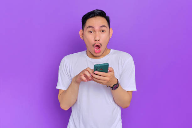 istock Shocked young Asian man in white t-shirt using smartphone and looking at camera with open mouth isolated on purple background 1428223928