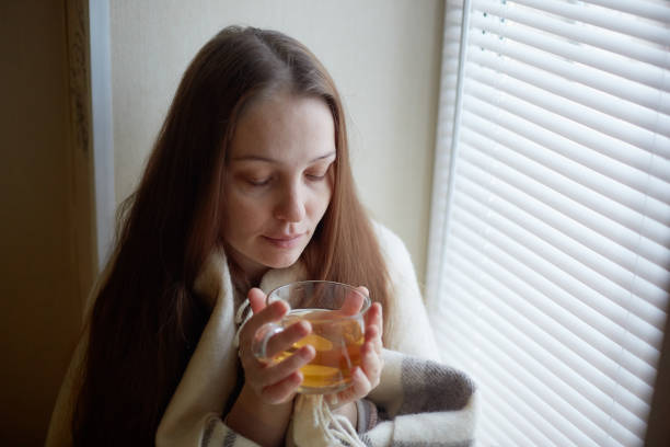 sick beautiful woman with colds and drinks hot tea with lemon sitting by the window in a country house in winter. stock photo