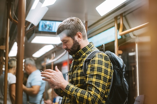 caucasian man in checkered plaid shirt with backpack traveling by metro, standing in subway car holding mobile telephone in hands.Image with selective focus.
