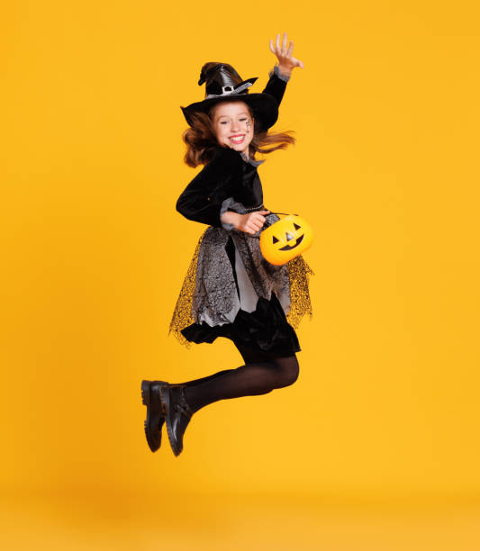 glückliches mädchen im hexenkostüm mit kürbiskorb springen und feiert halloween auf gelbem hintergrund - child jumping vegetable food stock-fotos und bilder