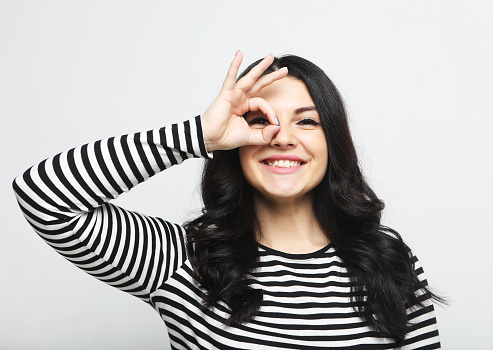 Cheerful beautiful adult woman lwearing striped shirt ooking at camera through hand finger binocular, laughing, having fun over white background