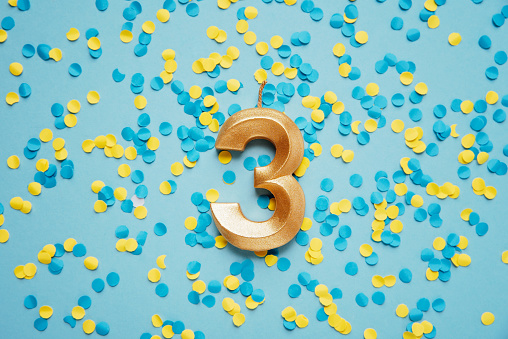 Number 3 three golden celebration birthday candle on yellow and blue confetti Background. three years birthday. concept of celebrating birthday, anniversary, important date, holiday