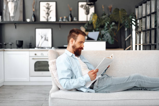 istock Young bearded handsome man reading book on couch at home 1428247847