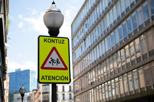 Triangular warning sign on a street light telling drivers to pay attention, written in Spanish and Basque, photographed in Bilbao, Spain, in summer
