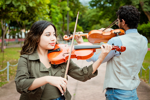 Two young violinists standing playing violin in a park. Portrait of man and woman together playing violin in park. Violinist man and woman back to back playing violin in a park outdoors