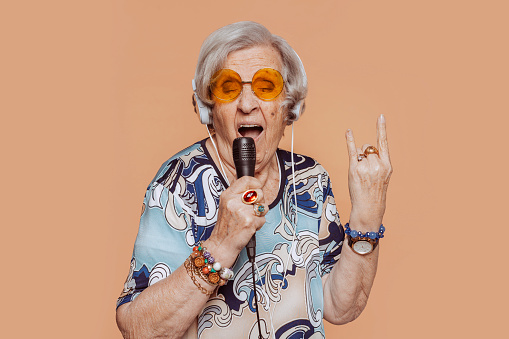 Funny elder grandmother portrait doing horns sign gesture with fingers while singing karaoke using microphone and headphones at studio isolated over beige background. Senior old woman rockstar granny.