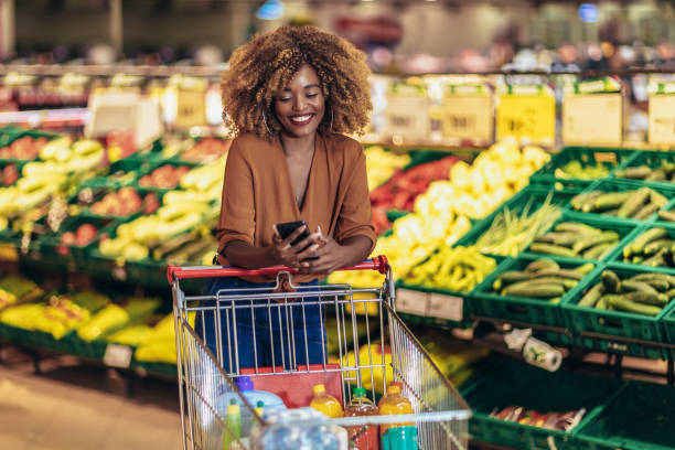Cheerful African American Woman In Supermarket Choosing Fresh Grocery Using Phone Cheerful African American Woman In Supermarket Choosing Fresh Grocery Using Phone groceries stock pictures, royalty-free photos & images