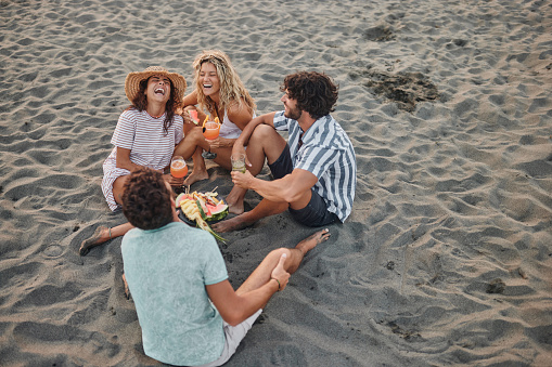 High angle view of group of cheerful friends communicating while eating fresh fruit during summer day in sand on the beach. Copy space.