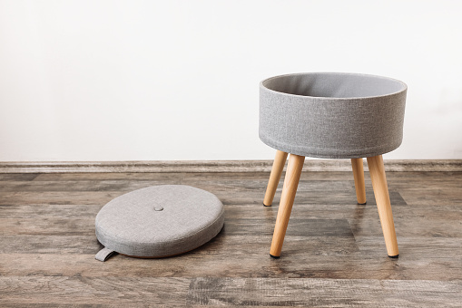 Trendy stool with and build-in storage space. Side view, white wall, copy space. Modern mulrifunctiional chair with wooden legs and removable lid. Round gray linen pouf