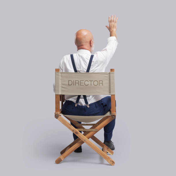 Professional filmmaker sitting on the director's chair and raising a hand, he is guiding the actors and the crew, back view