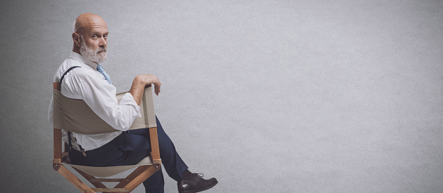 Confident film director sitting on the director's chair and looking at camera: video production, filmmaking and film industry concept