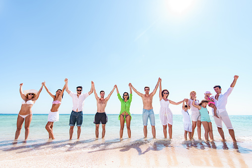 Large group of carefree people having fun while holding hands high up during summer day on the beach. Copy space.