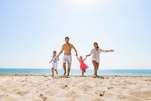 Young playful parents holding hands with their small kids and running on the beach. Copy space.