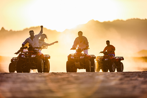 Group of carefree people driving quad bikes in the desert at sunset. Copy space.