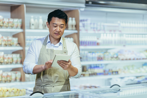 Internship at work. A young man, an Asian stager, an assistant works in a supermarket. Standing in an apron in the dairy department near the refrigerators, writing down the product in a notebook.