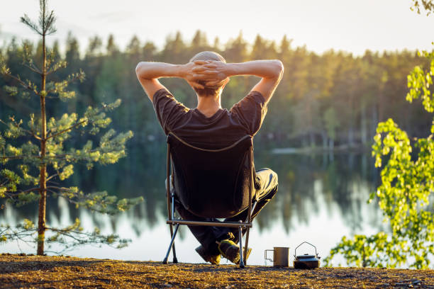 l'uomo è seduto su una sedia da campeggio sullo sfondo di un lago forestale in una bella sera d'estate. - riposarsi foto e immagini stock