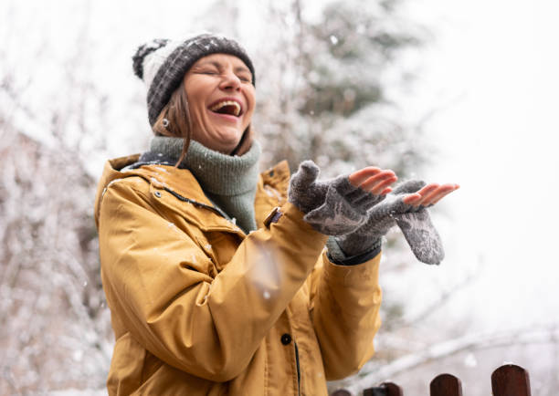 счастливая молодая женщина в желтом пальто и шляпе наслаждается падающим снегом и зимней погодой, ловя снежинки руками - mature women portrait hat human face стоковые фото и изображения