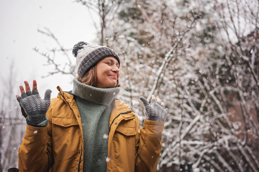 Happy young woman in yellow coat and hat enjoying falling snow and winter weather