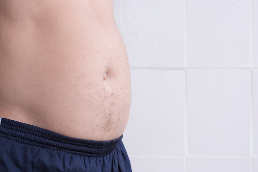 Midsection of fat adult man with excessive belly fat on white tile wall background, side view with copy space