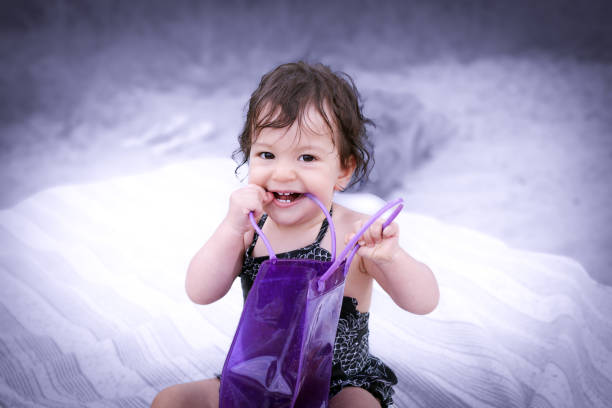Cute teething baby girl with brown hair and eyes in the swimsuit smiling and chewing on a purple plastic bag handle while sitting outside on a blanket in the beach on a blurry lilac background stock photo