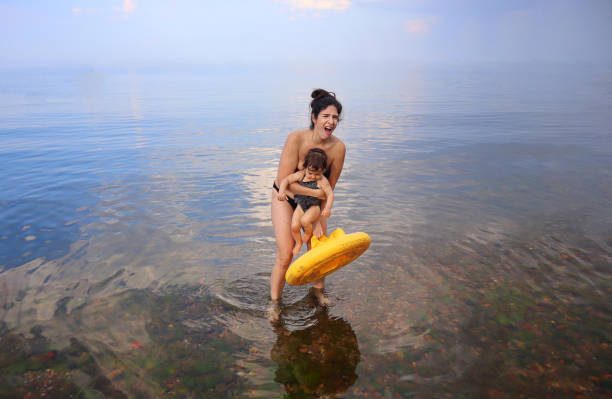 Mother in bikini holding yellow floaty and carrying baby daughter in her arms while making grimace and struggling to walk on a calm but shallow and rocky sea floor full of pebbles that hurt her feet stock photo