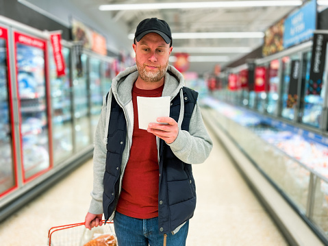 Color image depicting a mid adult male in the supermarket. He is looking at his shopping list while holding a basket of groceries. Focus is on the man with the supermarket aisle and products on the shelves totally defocused in the background.