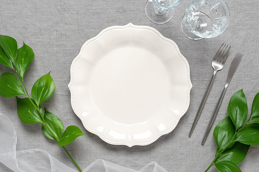Wedding table setting minimal. Empty beige plate mockup, cutlery, glasses, green leaves, gray linen textile background. Top view, flat lay.