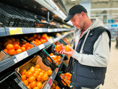 Color image depicting a man in his 30s looking at oranges in the fruit and vegetables aisle of a supermarket. Focus on the man with the rest of the aisle defocused.