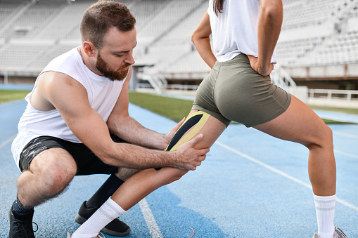 Male Coach Sticking Elastic Therapeutic Tape On Thigh Of Female Athlete To Ease Her Pain