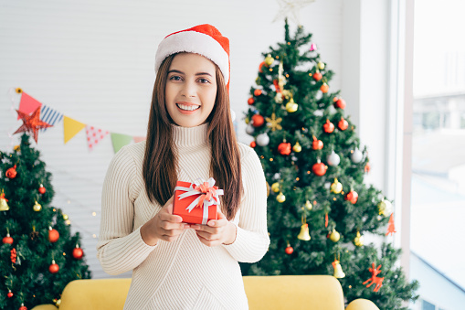 Young Asian woman wearing a sweater and Santa Claus hat smiles and holds a Christmas gift in a red box with ribbon in a living room with a decorated Christmas tree in the background.