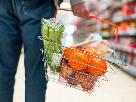 Low angle close up color image depicting a man holding a shopping basked filled with fresh groceries in the store. The products in the basket in include oranges, celery and nectarines.